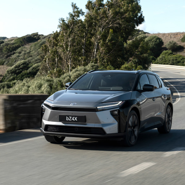 Pilkas modernus elektrinis visureigis „Toyota bZ4X“ važiuoja vingiuotu keliu, apsuptu žalumos ir medžių.