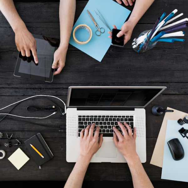 Asmuo dirba prie medinio stalo su nešiojamuoju kompiuteriu, šalia puodelis kavos, salotų dubenėlis, išmanusis telefonas, užrašų knygelės ir stalinė lempa prie lango.