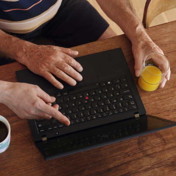 Du asmenys sėdi prie medinio stalo, vienas ro  do į nešiojamojo kompiuterio klaviatūrą, kitas laiko stiklinę apelsinų sulčių; ant stalo yra ir puodelis kavos.