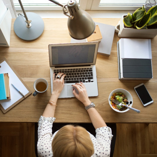 Asmuo dirba prie medinio stalo su nešiojamuoju kompiuteriu, šalia puodelis kavos, salotų dubenėlis, išmanusis telefonas, užrašų knygelės ir stalinė lempa prie lango.
