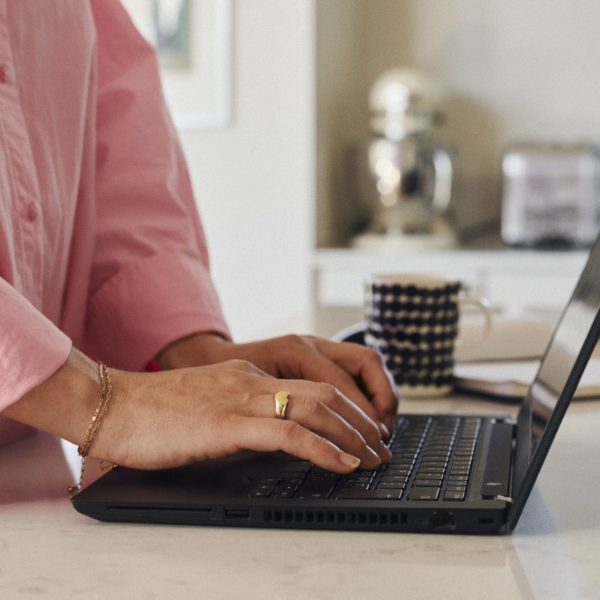 Asmuo rašo nešiojamuoju kompiuteriu, padėtu ant virtuvės stalo, fone matosi puodelis ir užrašų knygelė.