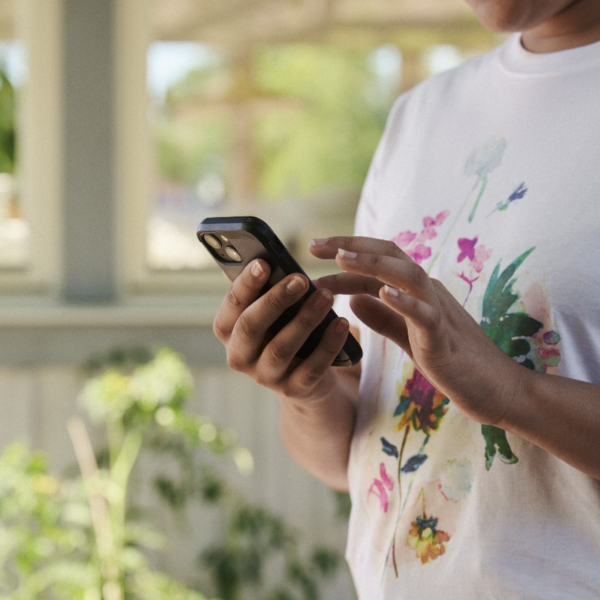 Asmuo dėvi baltus marškinėlius su spalvotu gėlių piešiniu ir laiko išmanųjį telefoną, žiūrėdamas į ekraną. Fonas – šviesi patalpa su augalais.