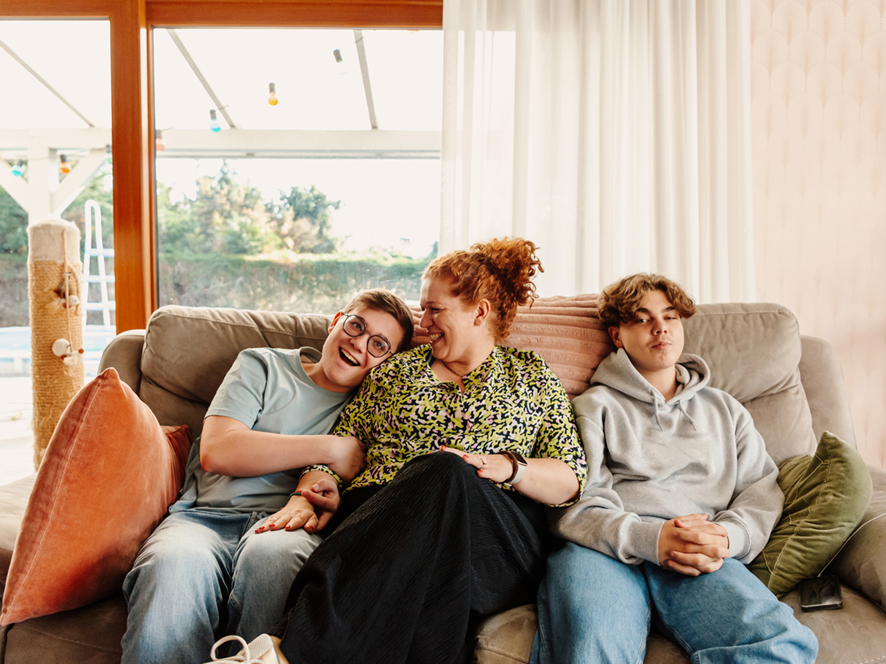 Three family members sitting closely together on a living room sofa, smiling and embracing, representing a sense of safety and comfort provided by life insurance