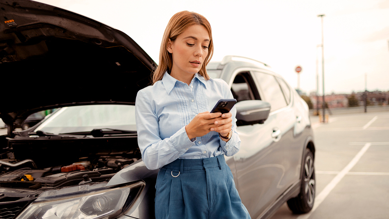 Moteris prie automobilio su pakeltu variklio dangčiu žiūri į telefoną.